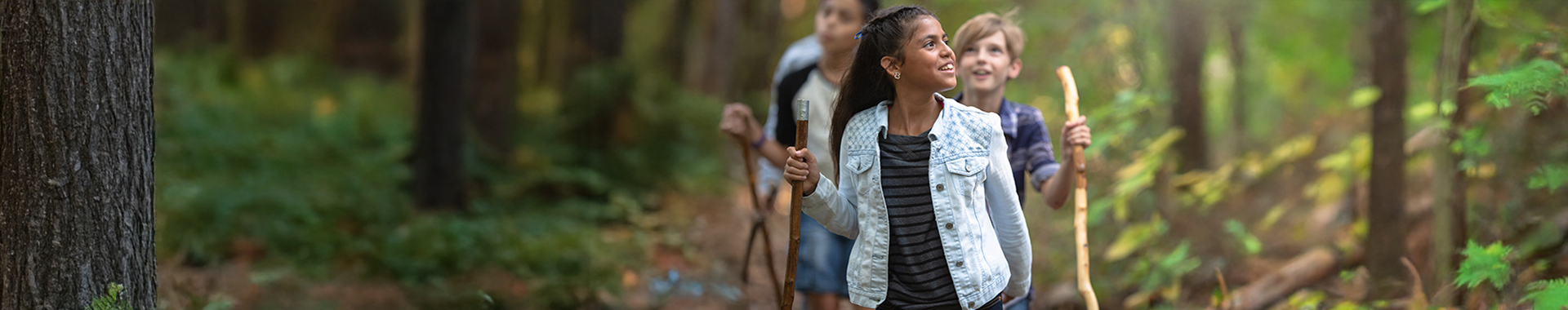 Three children hiking through a lush forest, with a cheerful girl leading the group and smiling.