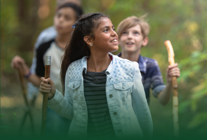 Three children hiking through a lush forest, with a cheerful girl leading the group and smiling.